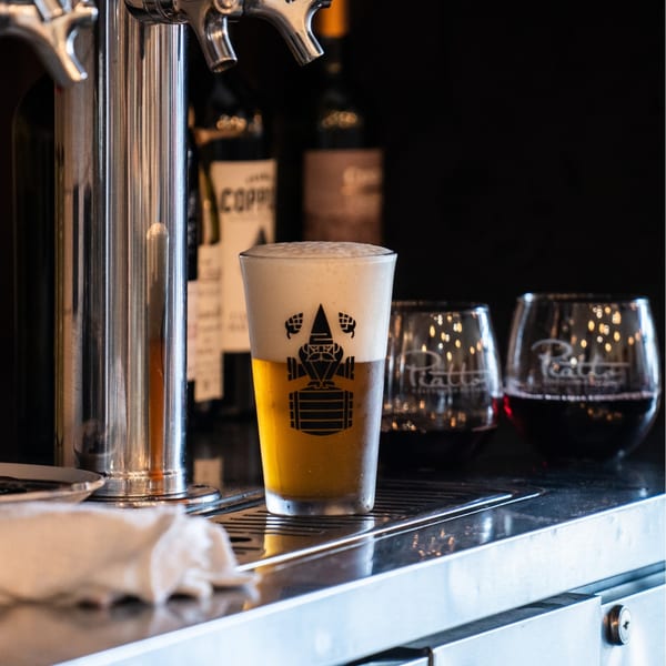A Piatto branded glass of beer with foam head, next to beer taps and wine glasses.