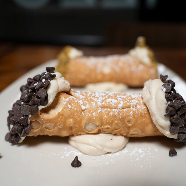 Two cannolis, one in focus, filled with cream, topped with chocolate chips and powdered sugar.